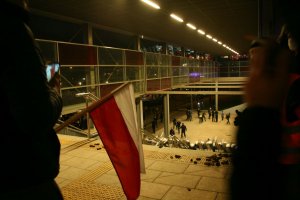 człowiek trzyma w ręku flagę polski stojąc na jednej z ulic. W tle barierki wiaduktu