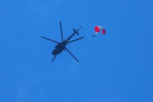 Policjant BOA skoczył ze śmigłowca na spadochronie. Widać helikopter i człowieka na spadochronie na tle nieba. Zdjęcie zrobione od dołu przez fotografa stojącego na ziemi.