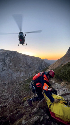 helikopter leci wśród szczytów górskich poniżej ratownik zajmuje się poszkodowanym turystą