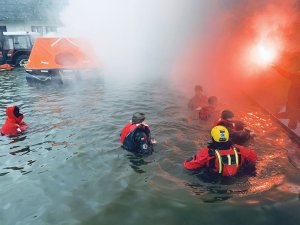 sanitariusze zanurzeni do połowy w wodzie w tle traktor, namiot ponton i na brzegu osoba z płonącą racą