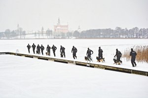 Policjanci z KMP w Suwałkach trasę przebiegli z psami służbowymi nad jeziorem Wigry. Jezioro zamarznięte, policjanci biegną z psami a w tle widać klasztor