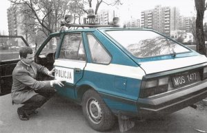 Radiowóz polonez 1989 rok produkcji silnik 1500 zwany noskiem ma koguty na dachu z napisem milicja. przy drzwiach klęczy policjant i przykleja na tylnych drzwiach napis policja.