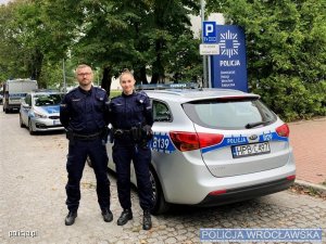Sierżant sztabowy Dariusz Wołkowski oraz sierżant Natalia Chołuj stoją na ulicy przy radiowozie