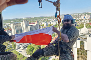 człowiek podczepiony liną na zewnątrz lecącego śmigłowca trzyma w rękach flagę Polski. W tle miasto