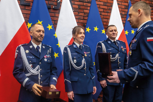 policjanci inspektor wręcza nadinsp. podziękowanie za służbę w tle flagi polski i unijne