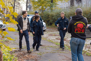 zdjęcia z planu filmopwego. policjant i aktirzy stoją przy radiowozie. W tle operator kamery