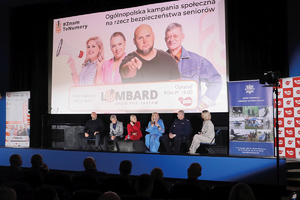 wielki bilbord z wyświetloną prezntacją a na scenie siedzi kilka osób i dyskutuje