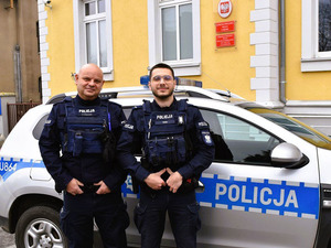Dwaj policjancie przy swoim radiowozie, w tle budynek komendy.