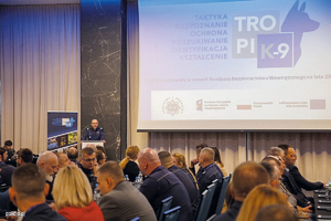 konferencja. Logo projektu wyświetlone na ścianie. Siedzą ludzie a przy mównicy jeden z policjantów