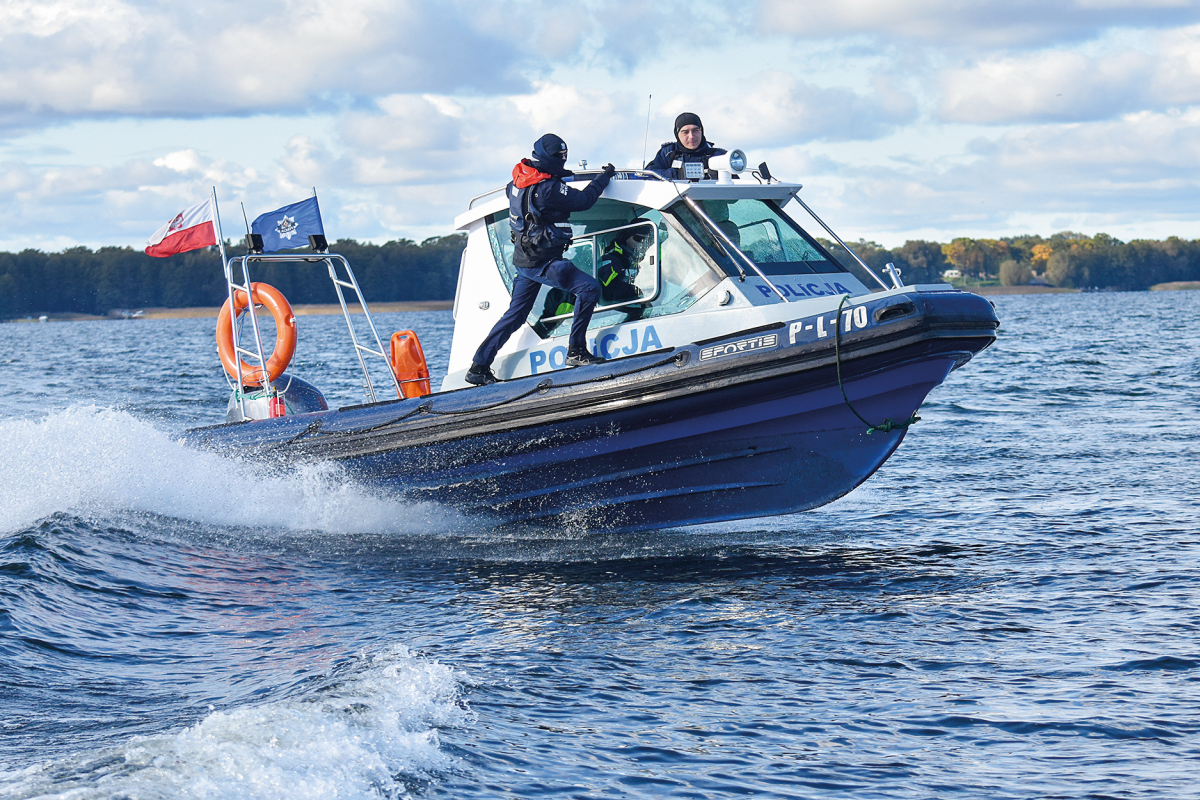 łódź patrolowa sunie z dużą prędkościa po falach. na niej dwóch policjantów po bokach burty